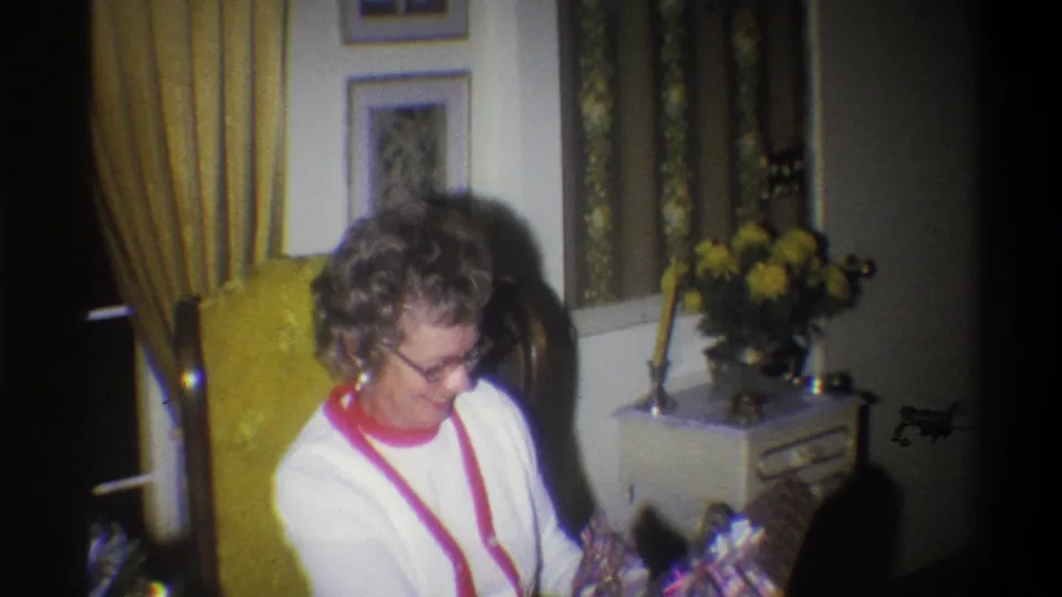 The woman looks down toward the birthday gift in a 1973 Troy, New York living room with yellow curtains, framed pictures, and flowers nearby.
