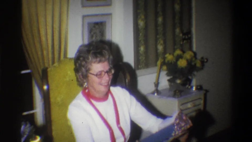 A seated woman smiles and extends the wrapped present in a warm 1973 Troy, New York home interior with curtains and flowers behind her.