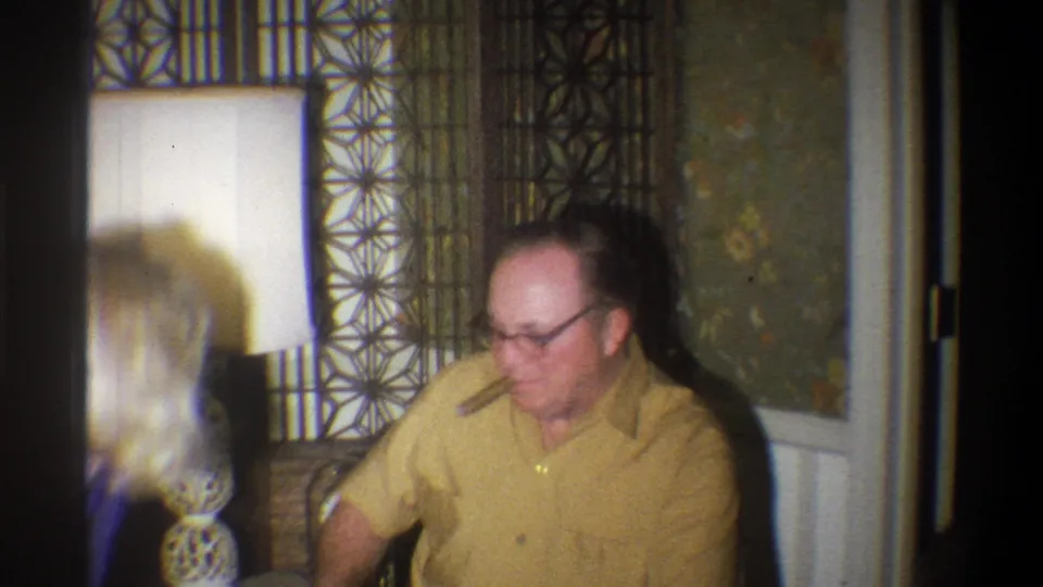A man in glasses and a yellow shirt sits in a 1973 Troy, New York home interior near geometric wall decor and a decorative lamp.
