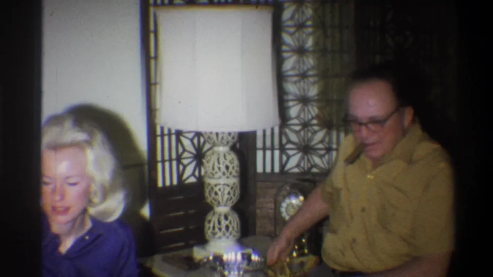 1973 Troy, New York frame with a blonde woman in blue seated beside a man in glasses in a dim living room.