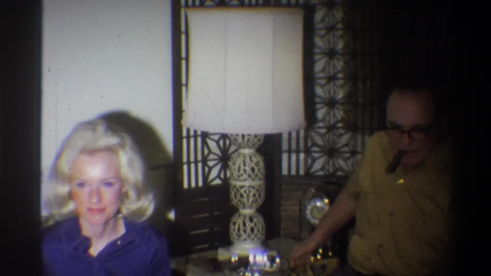 1973 Troy, New York home-movie frame showing a woman in blue near a lamp, with a man holding a cigar in shadow.