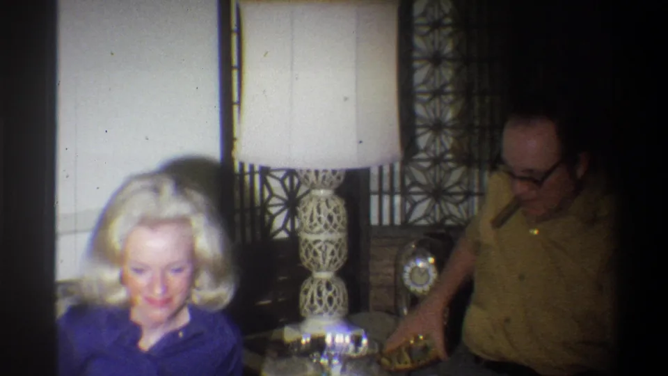 1973 Troy, New York frame of a woman looking down beside a man with a cigar near an ornate lamp and table.