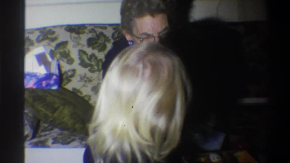 1973 Troy, New York frame with an older woman in glasses facing a blonde child beside a floral couch in a private interior.