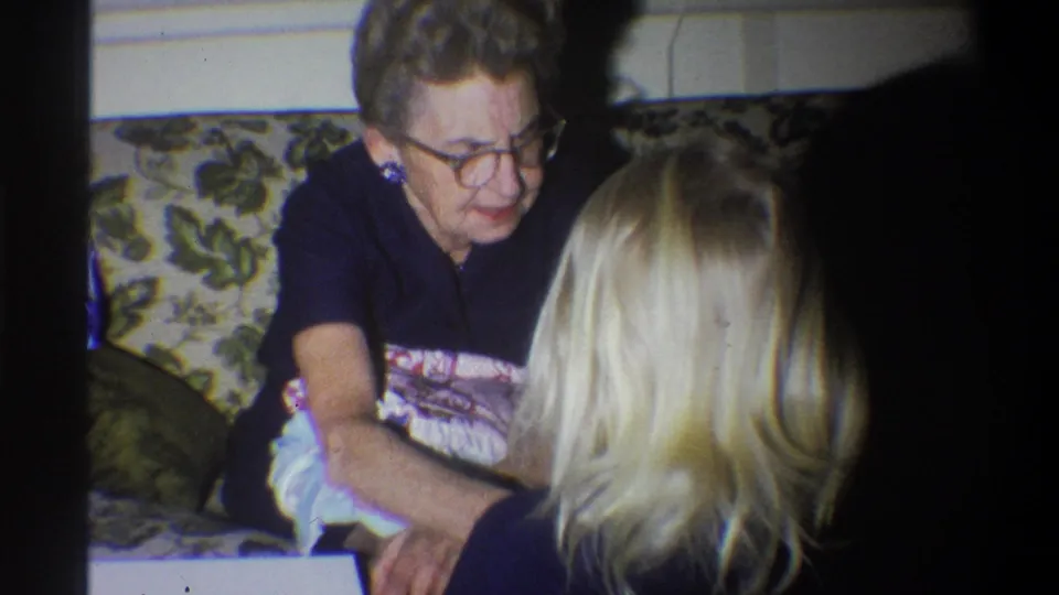 1973 Troy, New York home-movie frame showing an older woman with glasses holding a soft package or fabric as a blonde child faces her.