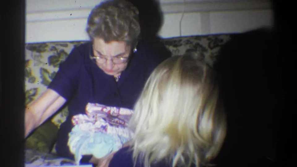 1973 Troy, New York frame of an older woman examining a soft package or fabric while a blonde child sits close in a living room.