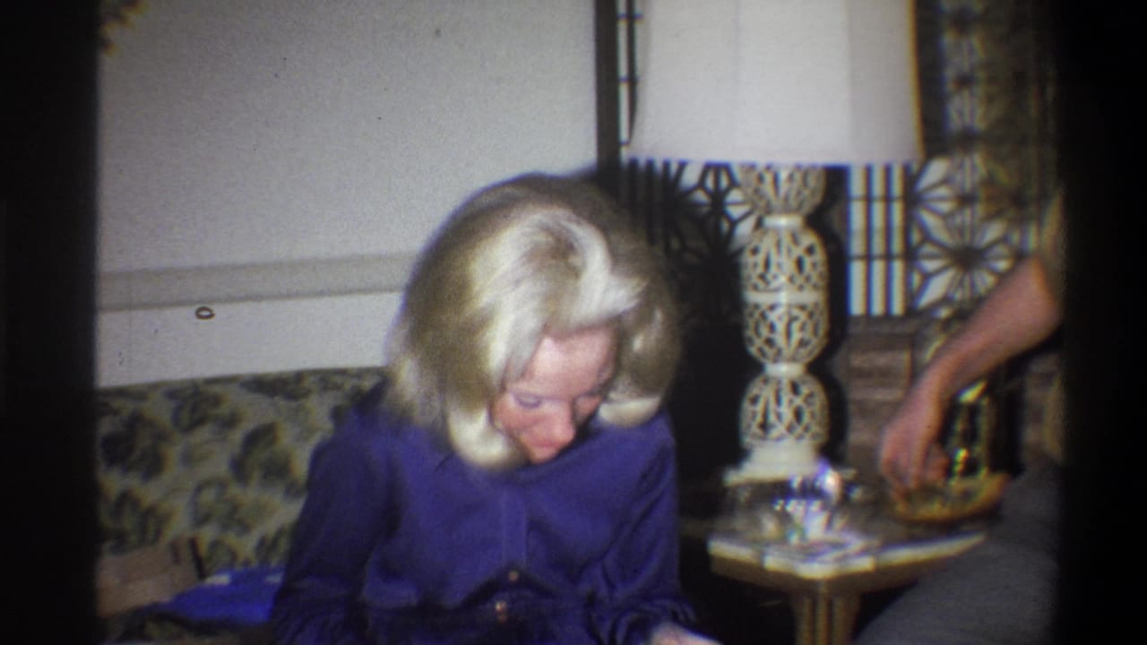 1973 Troy, New York home-movie frame showing a blonde woman in purple seated on a floral couch near a ceramic lamp.