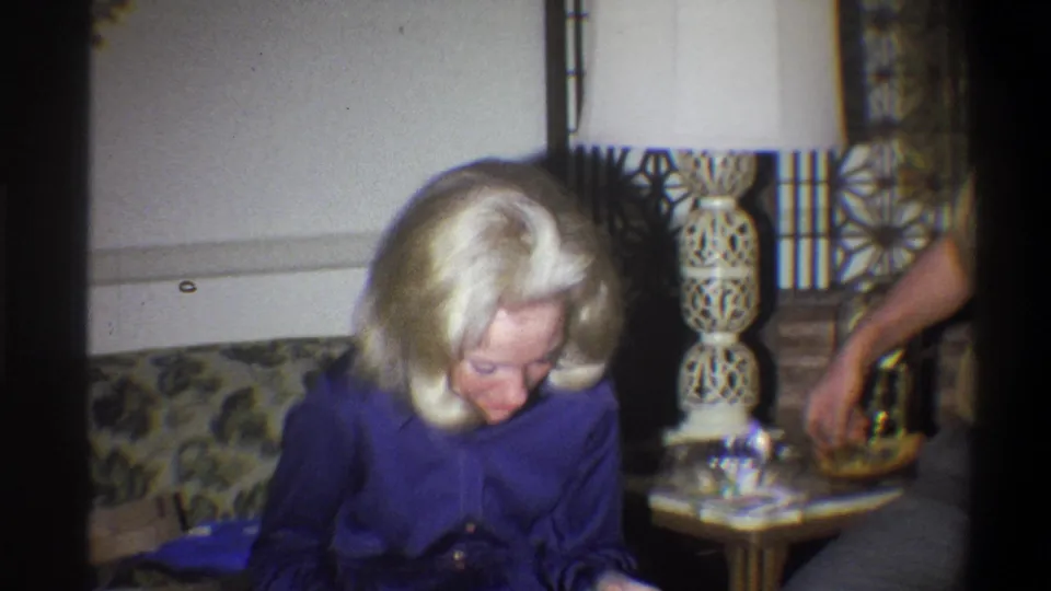 1973 Troy, New York home-movie frame showing a blonde woman in purple seated on a floral couch near a ceramic lamp.