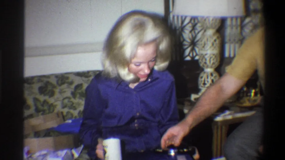 1973 Troy, New York home-movie frame showing a woman with a white cup while a man reaches near a living-room table.