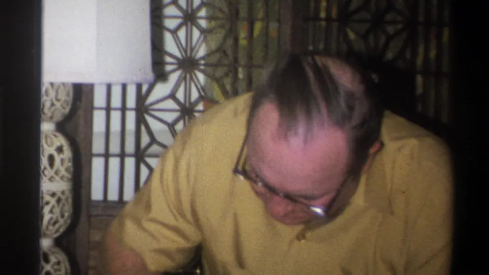 1973 Troy, New York frame showing a man in a yellow shirt and glasses leaning forward near a lamp and patterned divider.