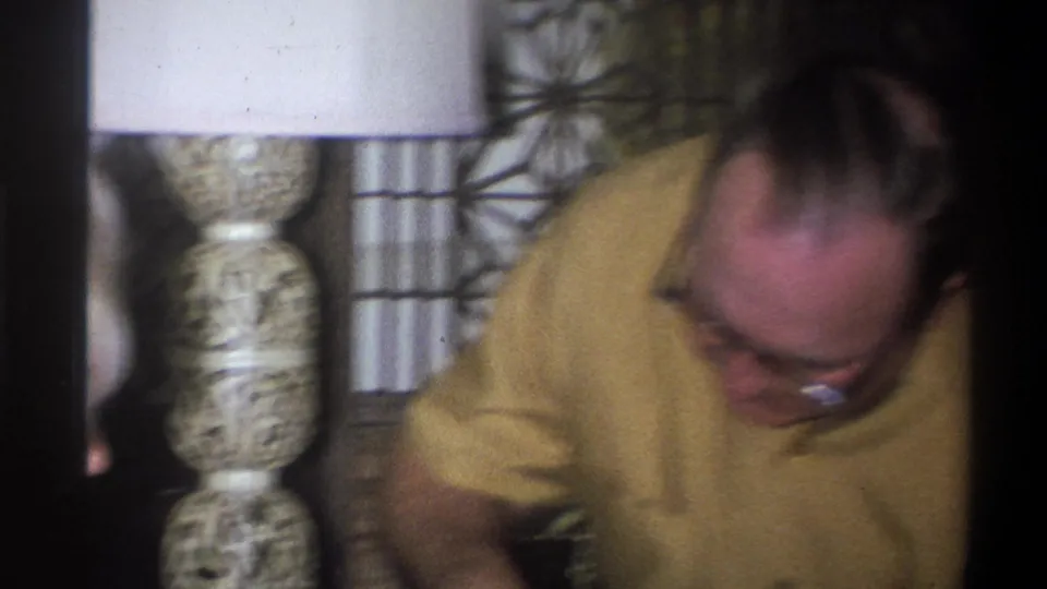A man in a yellow short-sleeved shirt and glasses leans forward in a 1973 Troy, New York home interior.