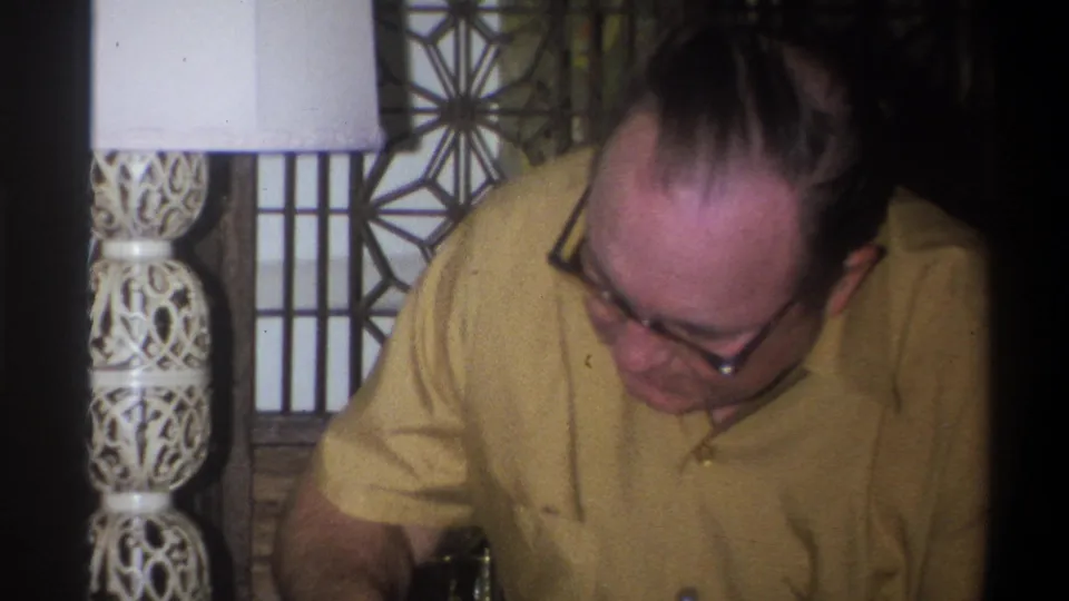 The man in a yellow shirt looks down near a table, with an ornate lamp and patterned interior panels behind him.