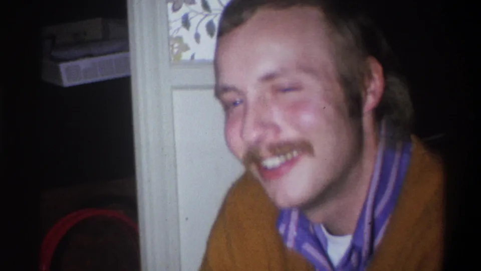 A smiling mustached man in an orange sweater and striped shirt appears in the same 1973 Troy, New York home-movie setting.