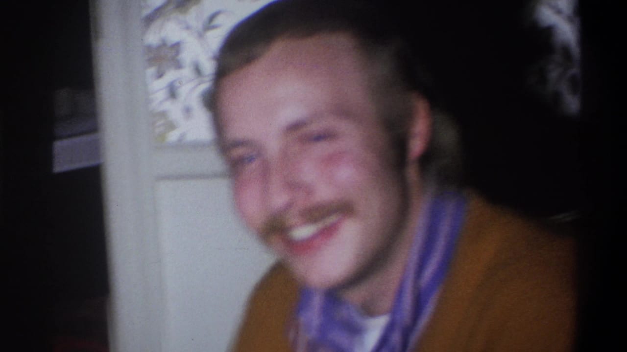 In this 1973 Troy, New York home-movie frame, a mustached man in a brown sweater laughs near a floral-patterned interior surface.