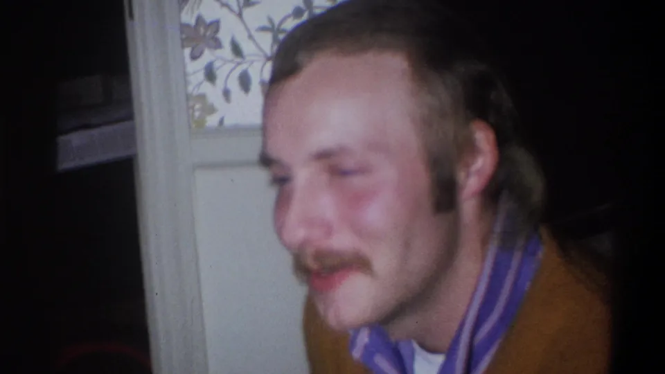 A 1973 Troy, New York frame shows the man smiling in partial profile, with his striped collar and brown sweater visible in dim light.
