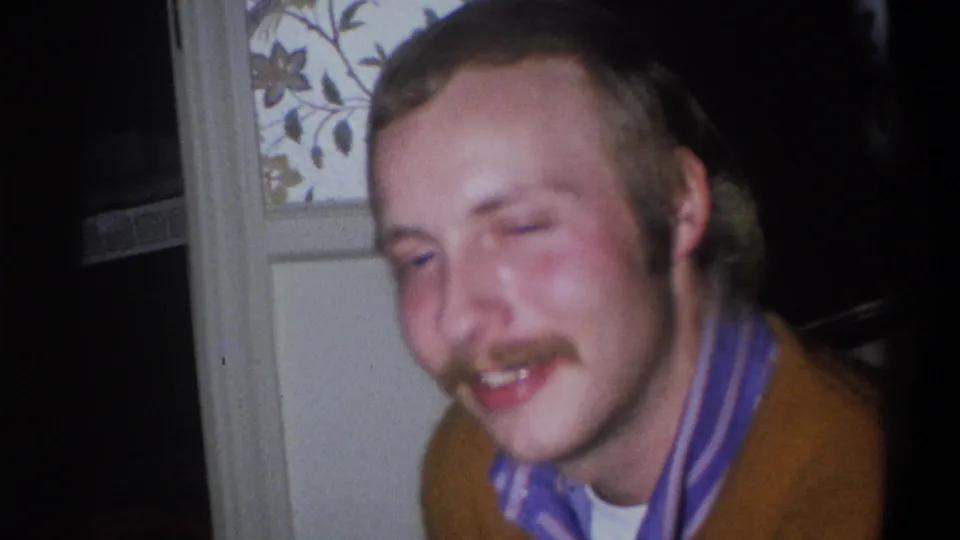 The 1973 home-movie frame shows the mustached man leaning forward, smiling with one eye partly closed beside a patterned interior surface.