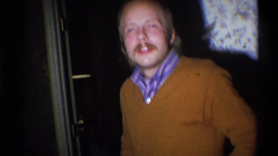 In this 1973 Troy, New York frame, the man faces the camera from farther back, wearing a brown sweater and striped collar in a dark interior.