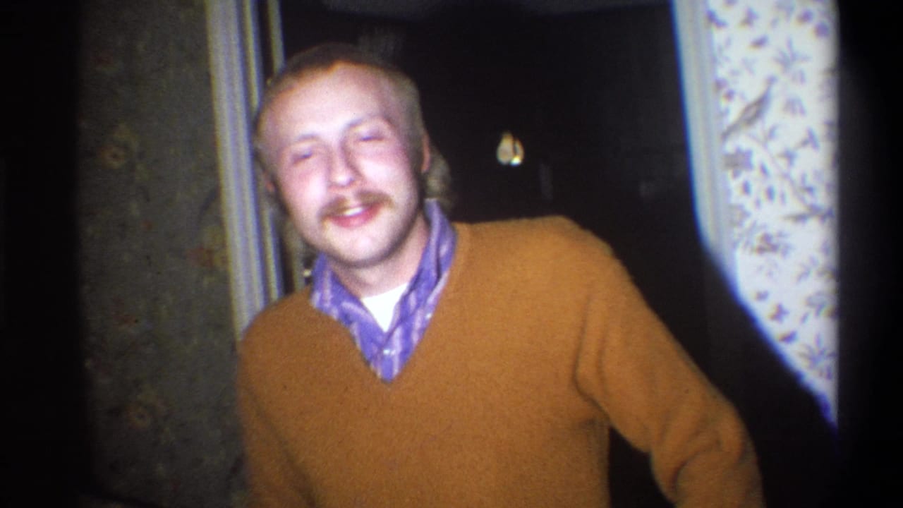 Mustached young man in a brown sweater smiling in a dim doorway.
