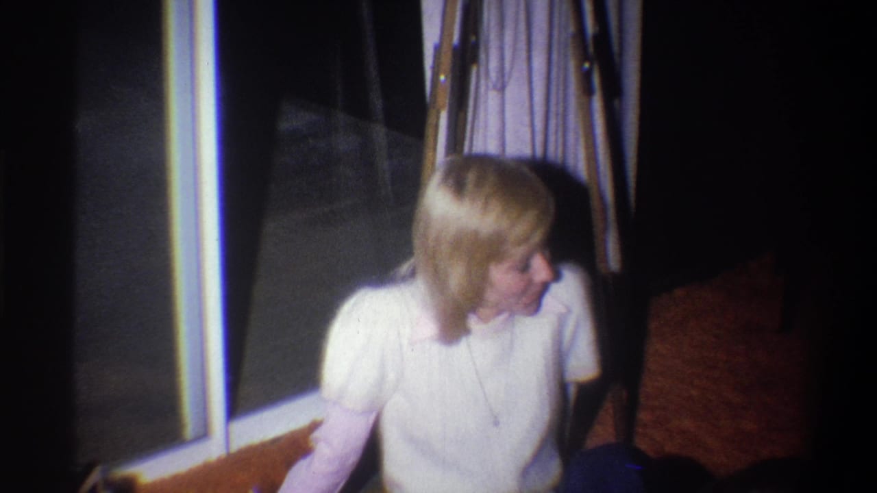 Blonde woman in a white sweater sits near a window in a dim living room.