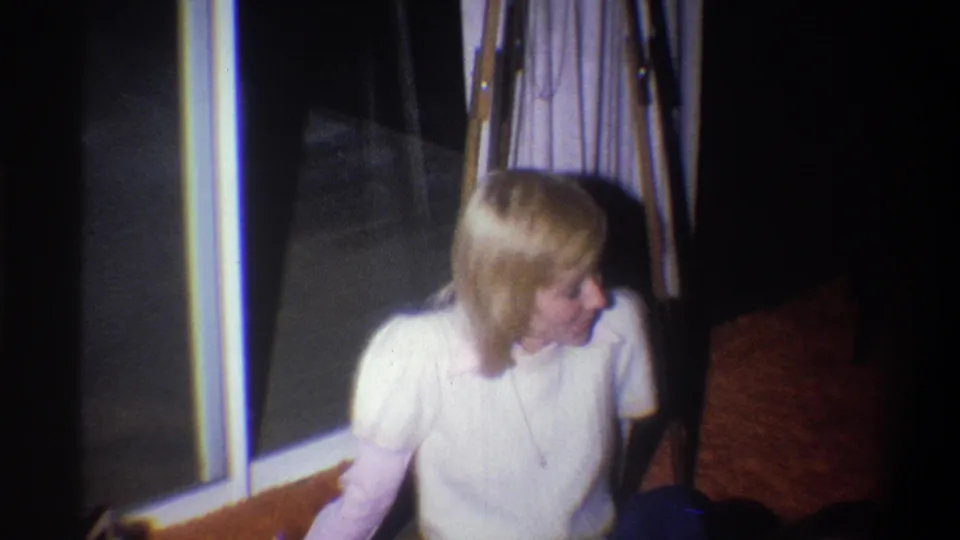 In a 1973 Troy, New York home-movie frame, a blonde woman in a white sweater sits on the floor near curtains and a window.