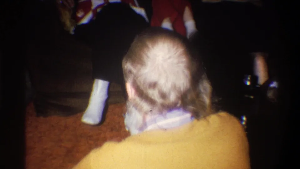 A mustached man in a mustard sweater is seen mostly from behind while seated among adults on a carpeted living-room floor.