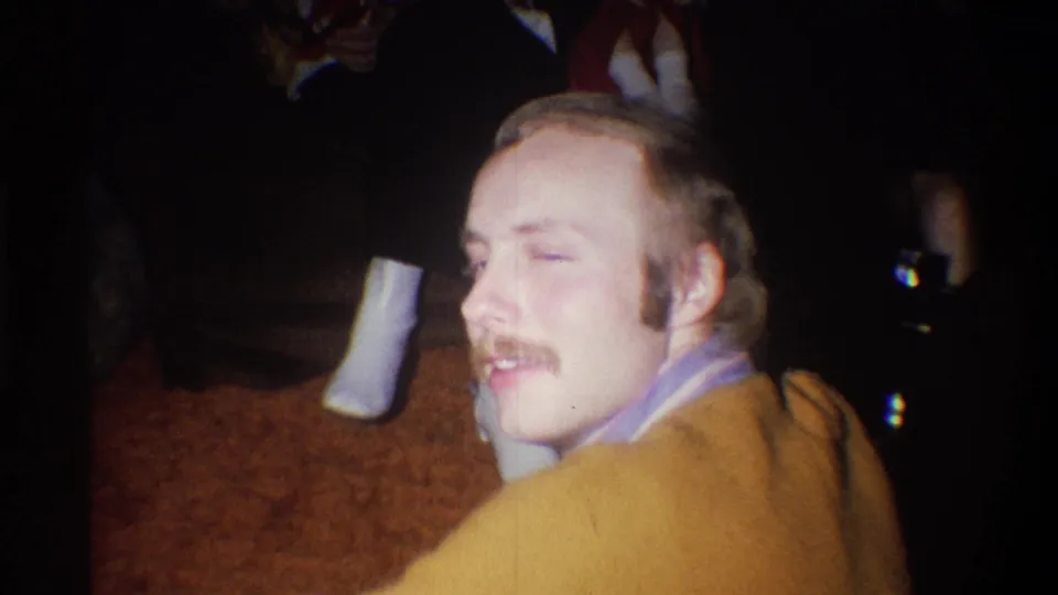 A mustached man with sideburns in a mustard sweater smiles toward the camera or another person in the 1973 Troy interior.