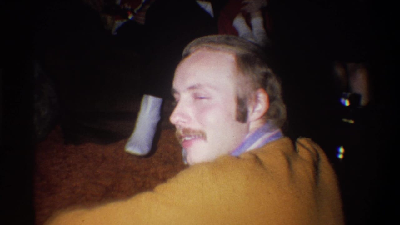 Mustached man in a mustard-orange sweatshirt turns toward the camera in a dim interior.