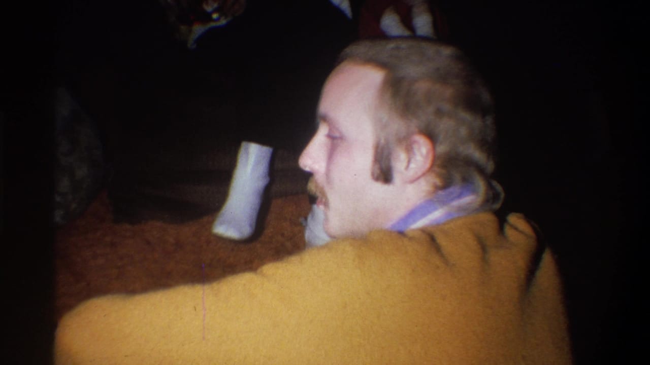 The 1973 Troy, New York frame shows the man in profile, seated or low in frame, with legs and white socks visible in the dark room behind him.