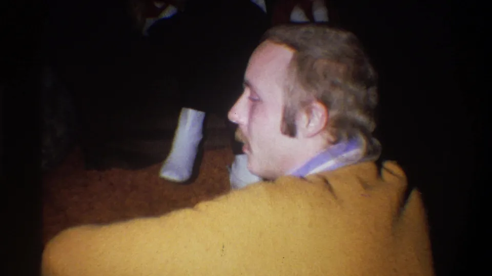 A close home-movie view shows the mustached man in profile inside a dim Troy, New York interior, with soft 8mm grain and muted color.