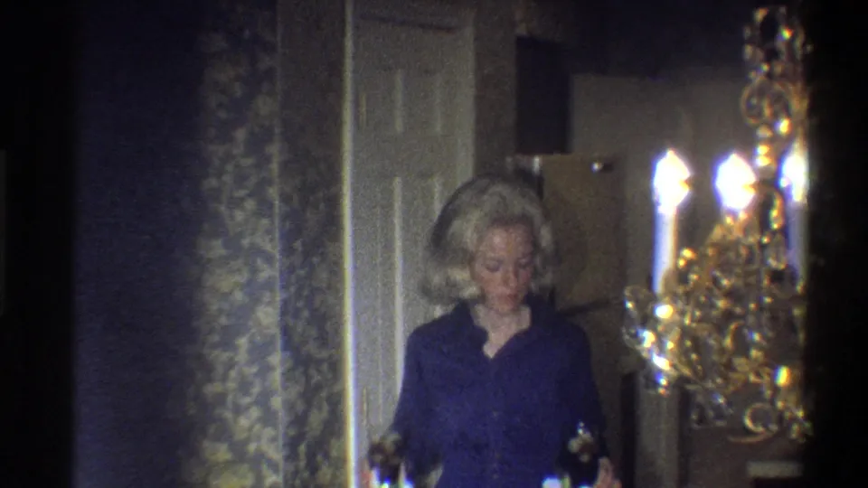 A blonde woman in a dark blue top stands in a warmly lit private room near patterned wallpaper, doorway trim, and a chandelier.