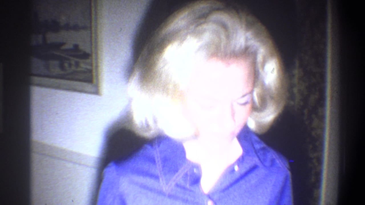 A blonde woman in a blue dress appears in a dim private interior in 1973 Troy, New York home-movie footage.