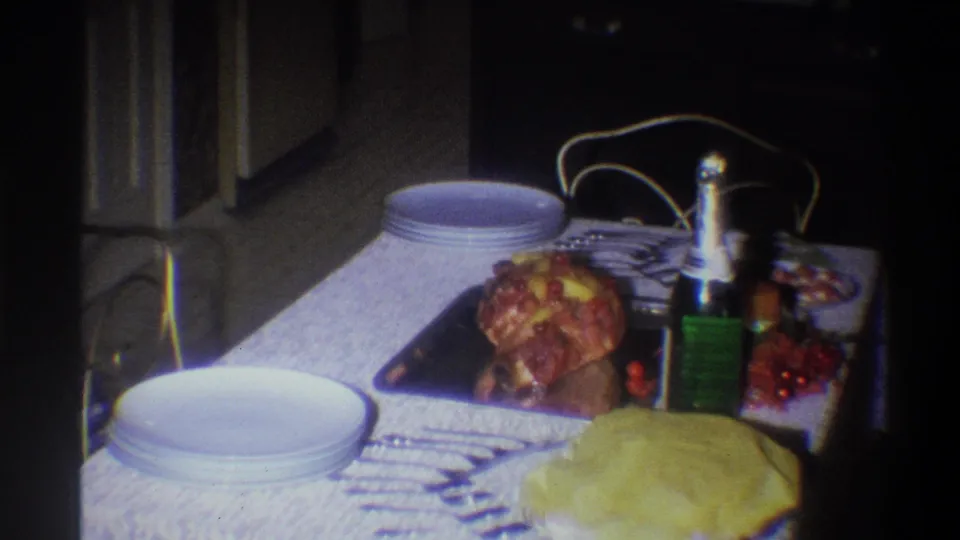 The home-movie camera frames the 1973 dinner table more closely, showing stacked plates, roasted chicken, a bottle, and a lace covering.