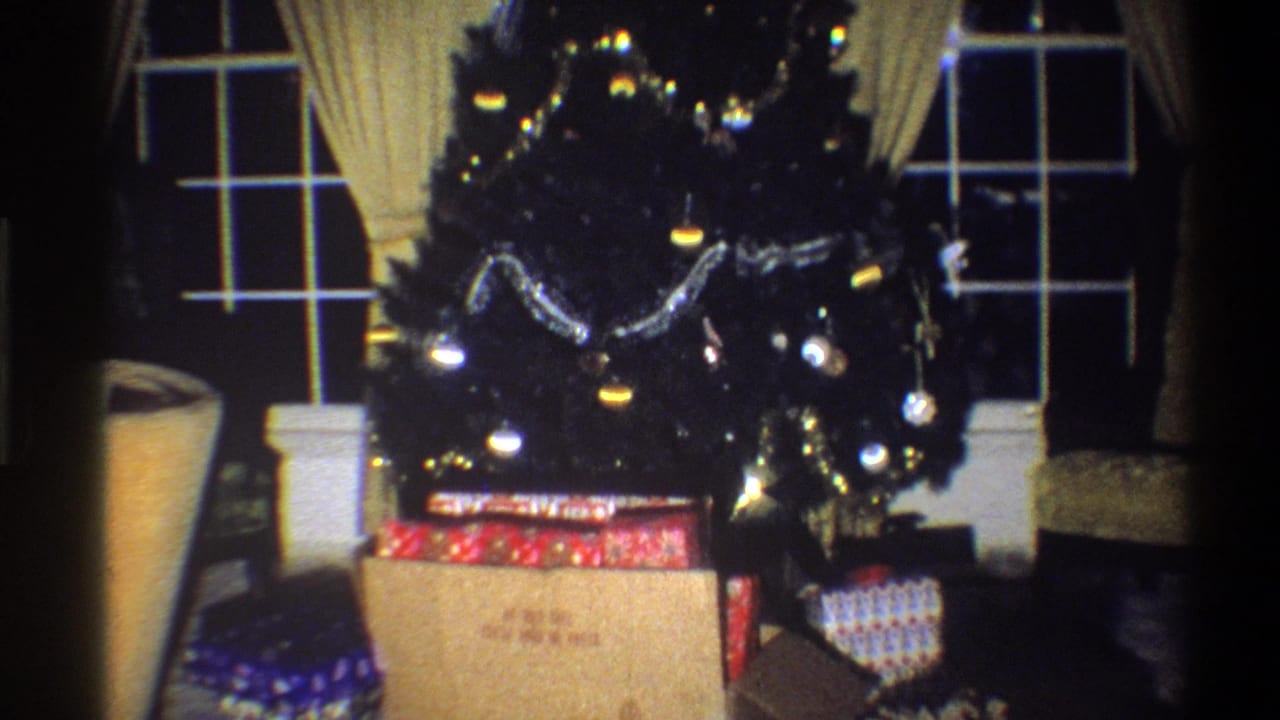 1973 Troy, New York home-movie frame showing a decorated Christmas tree, gifts, drapes, and windows in a dim living room.
