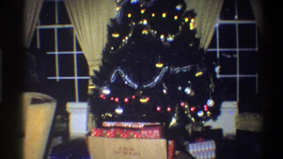1973 Troy, New York home-movie frame with a lit Christmas tree, garland, ornaments, and presents in a private living room.