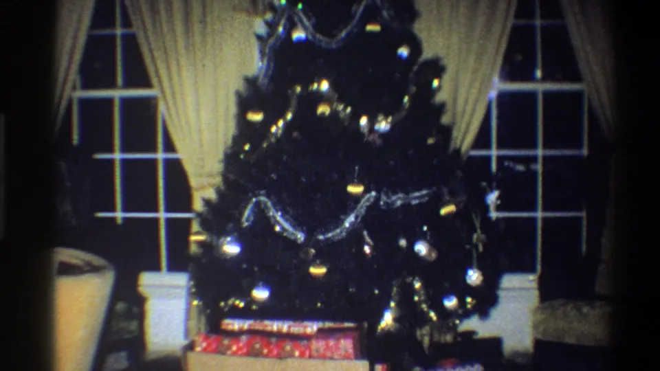 1973 Troy, New York frame showing a decorated Christmas tree, wrapped presents, yellow drapes, windows, and part of an armchair.