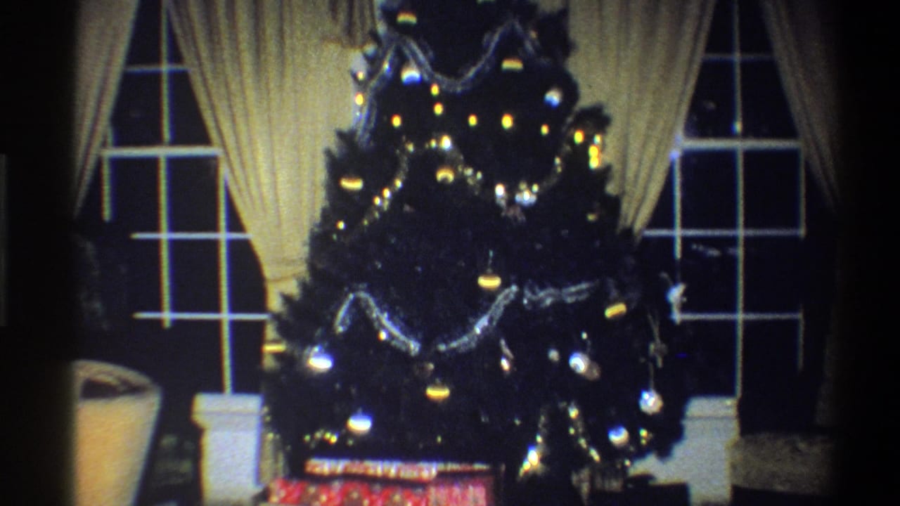 1973 Troy, New York home-movie frame showing a decorated Christmas tree between cream-draped windows with ornaments, tinsel, lights, and gifts.