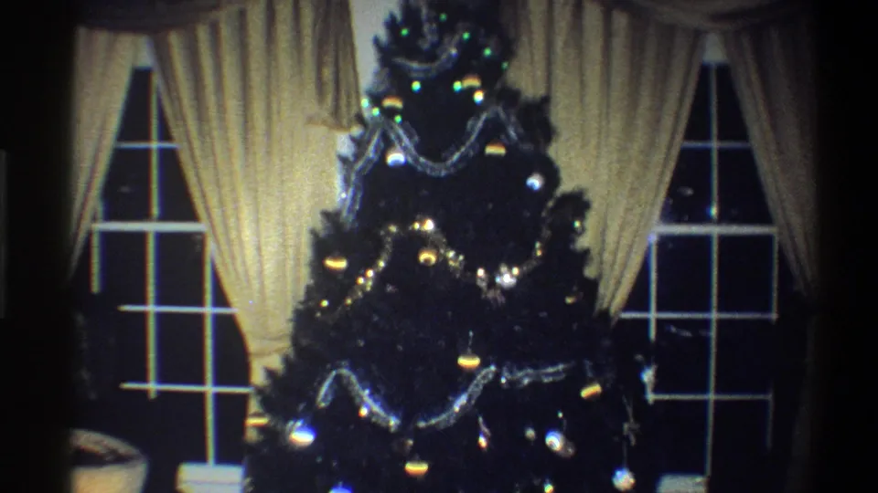 1973 Troy, New York frame of a Christmas tree with silver garlands, ornaments, and lights set between cream curtains and dark window panes.