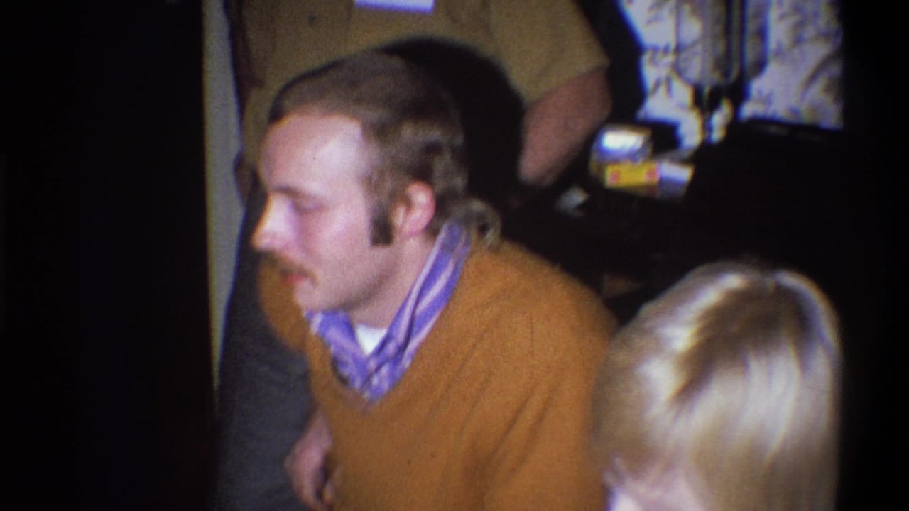 1973 Troy, New York home-movie frame with a mustached man in a brown sweater near other adults in a dim living room.