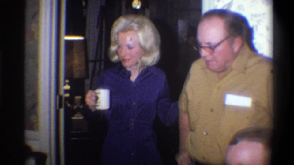 1973 Troy, New York home-movie frame of two adults standing close together during an informal indoor gathering.