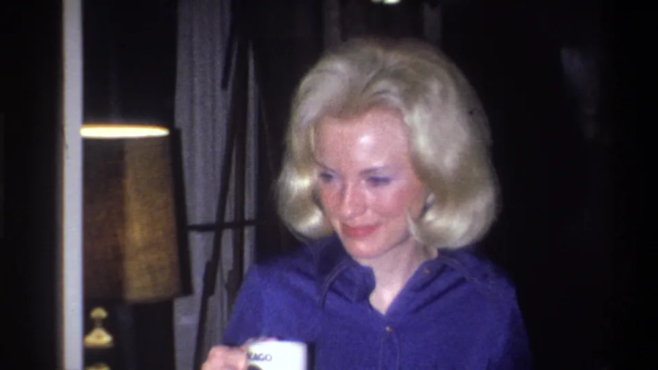 1973 Troy, New York home-movie frame with a blonde woman in blue holding a white mug beside curtains and a table lamp.
