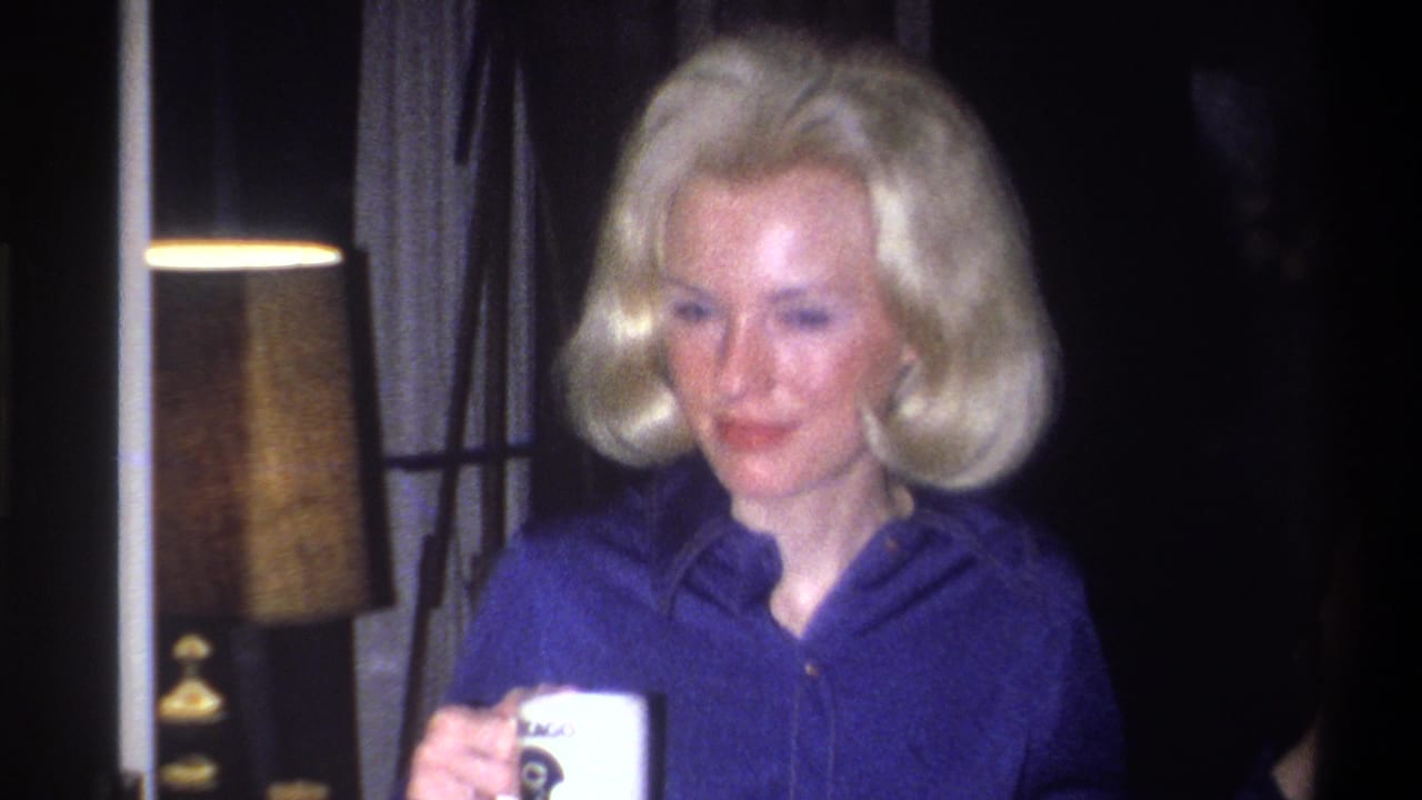 1973 Troy, New York home-movie frame of an adult blonde woman in a blue collared shirt holding a white cup in a dim interior.