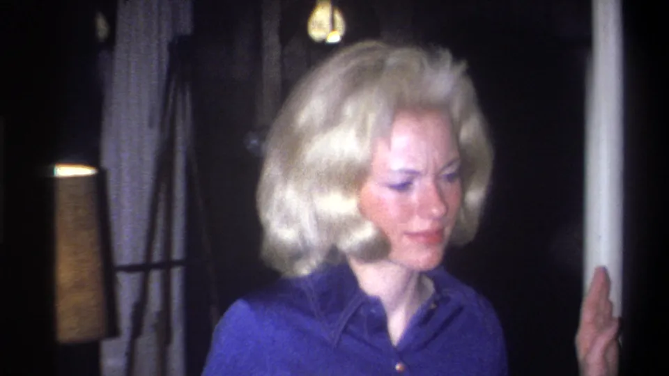 Adult woman with blonde bouffant hair turns away from the camera in a dim 1973 Troy interior with a lamp behind her.