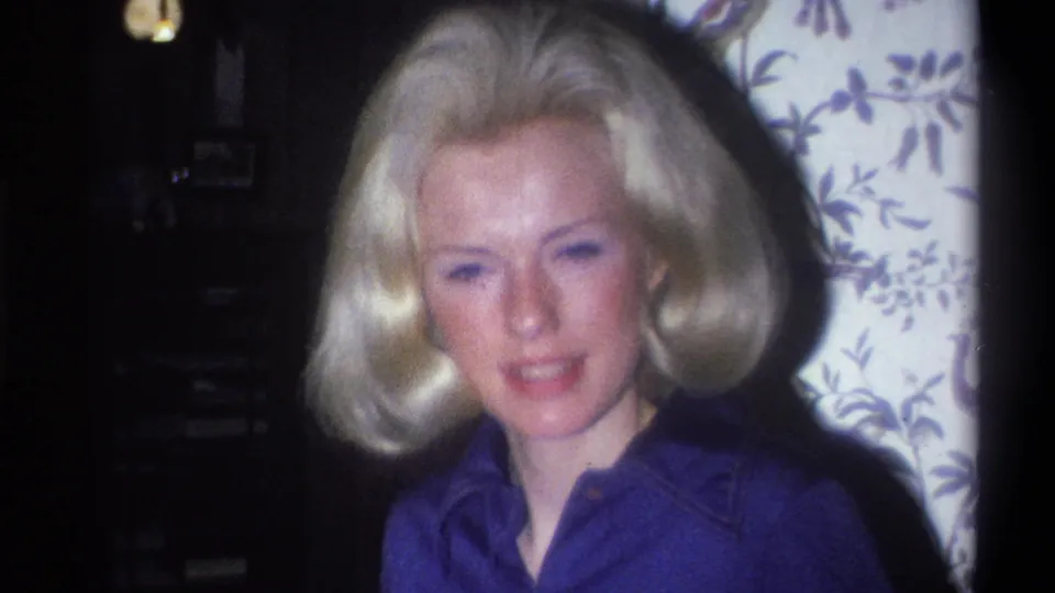 Close home-movie view of the blonde woman in a blue shirt facing the camera beside patterned wallpaper in Troy, New York.