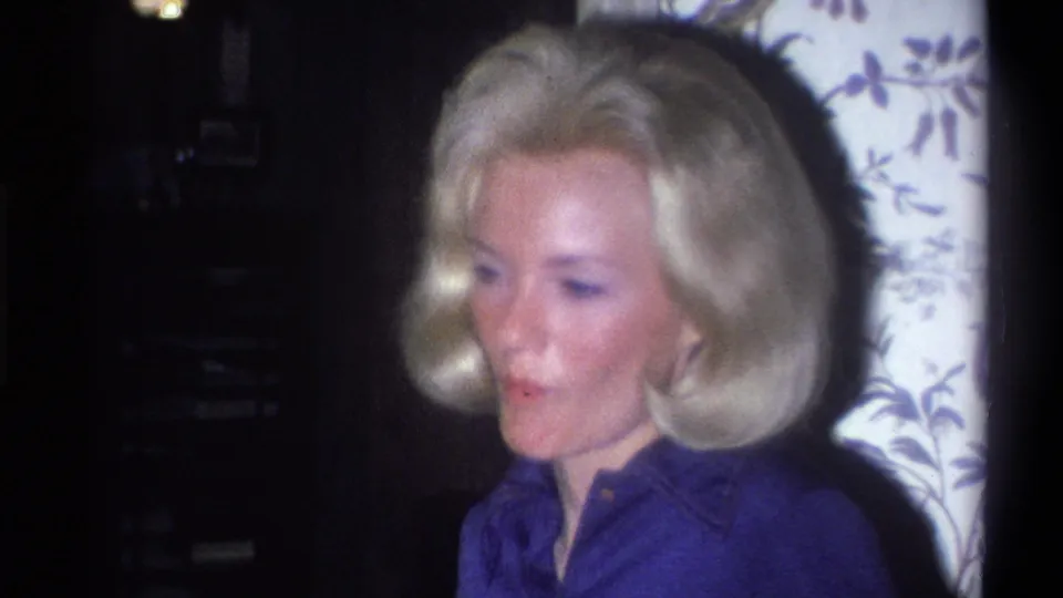 Adult woman in a blue collared shirt looks to the side in a soft 8mm home-movie frame with patterned wallpaper behind her.
