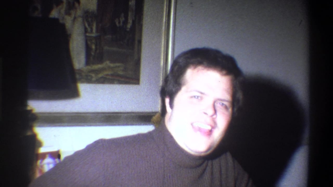 In a 1973 Troy, New York home-movie frame, an adult man in a dark turtleneck looks toward the camera inside a softly lit room.