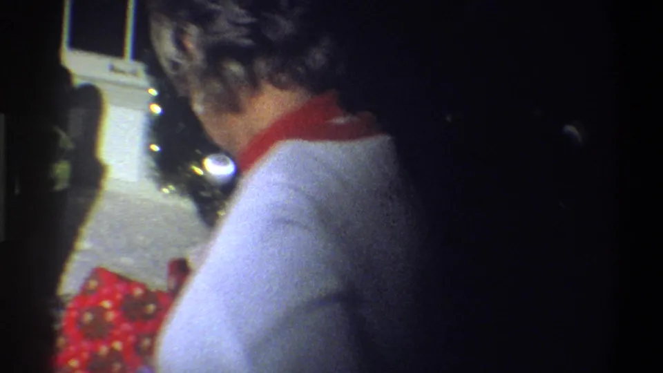 A woman in a white sweater with a red collar is seen from behind in a dim 1973 Troy, New York home interior.