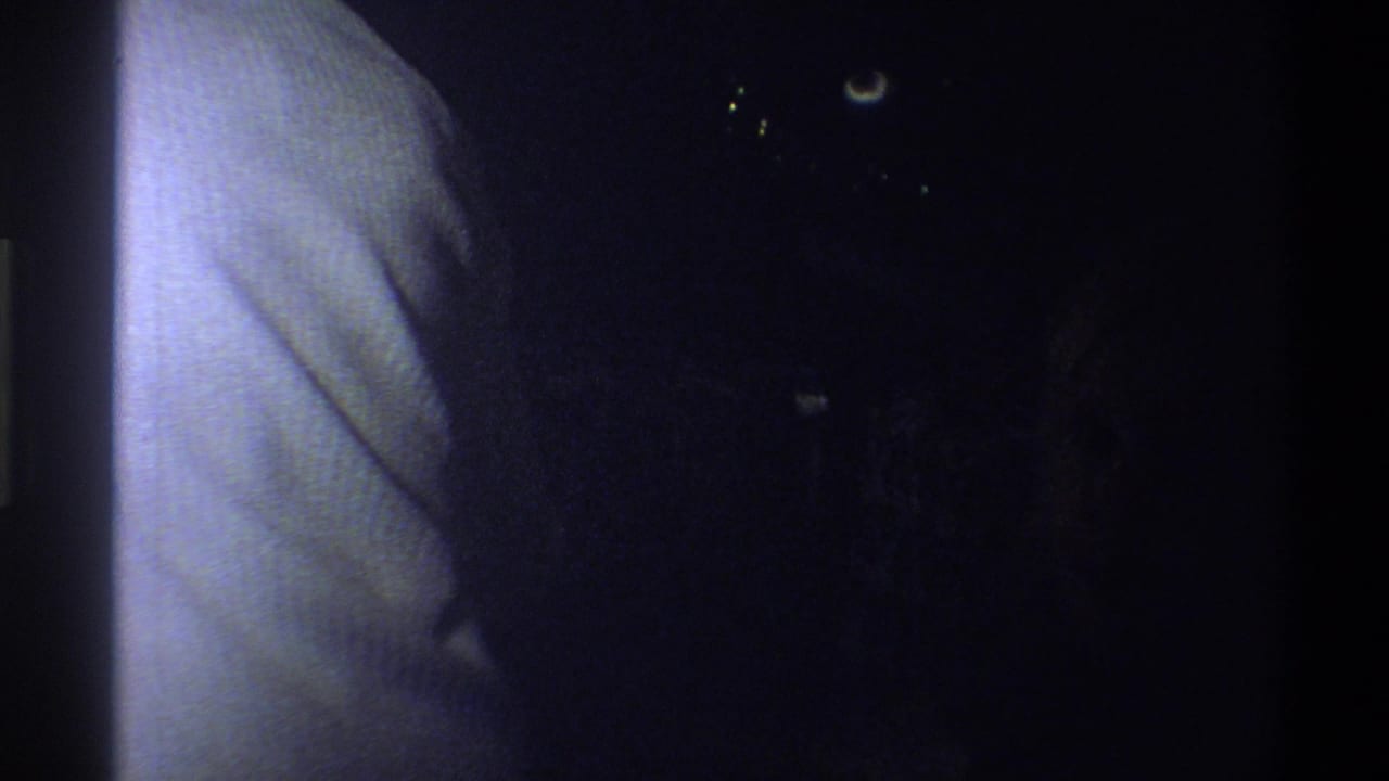 Dark 1973 8mm home-movie frame from Troy, New York, with a pale vertical surface and faint points of light in shadow.