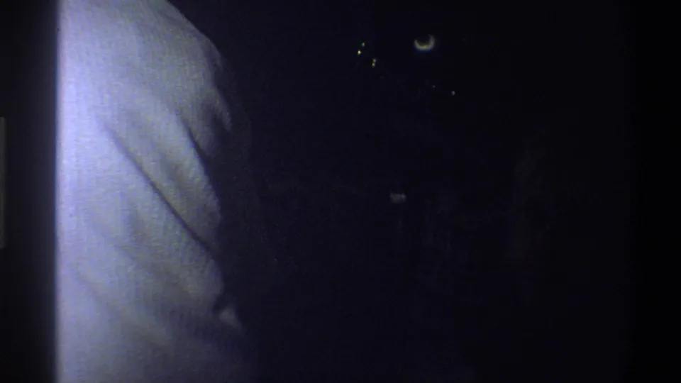 Dark 1973 8mm home-movie frame from Troy, New York, with a pale vertical surface and faint points of light in shadow.