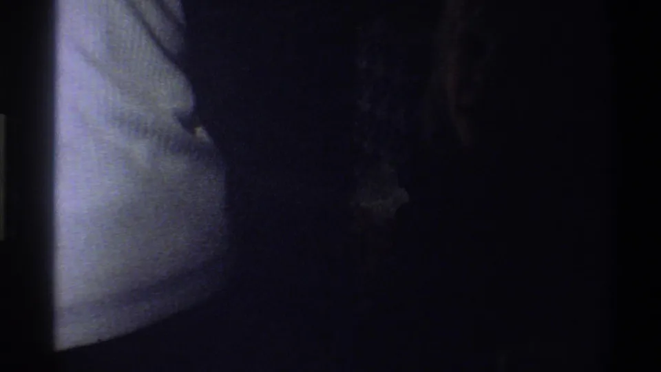 Very dark 8mm frame with a pale vertical edge and indistinct shapes, likely part of the transition before the indoor scene.