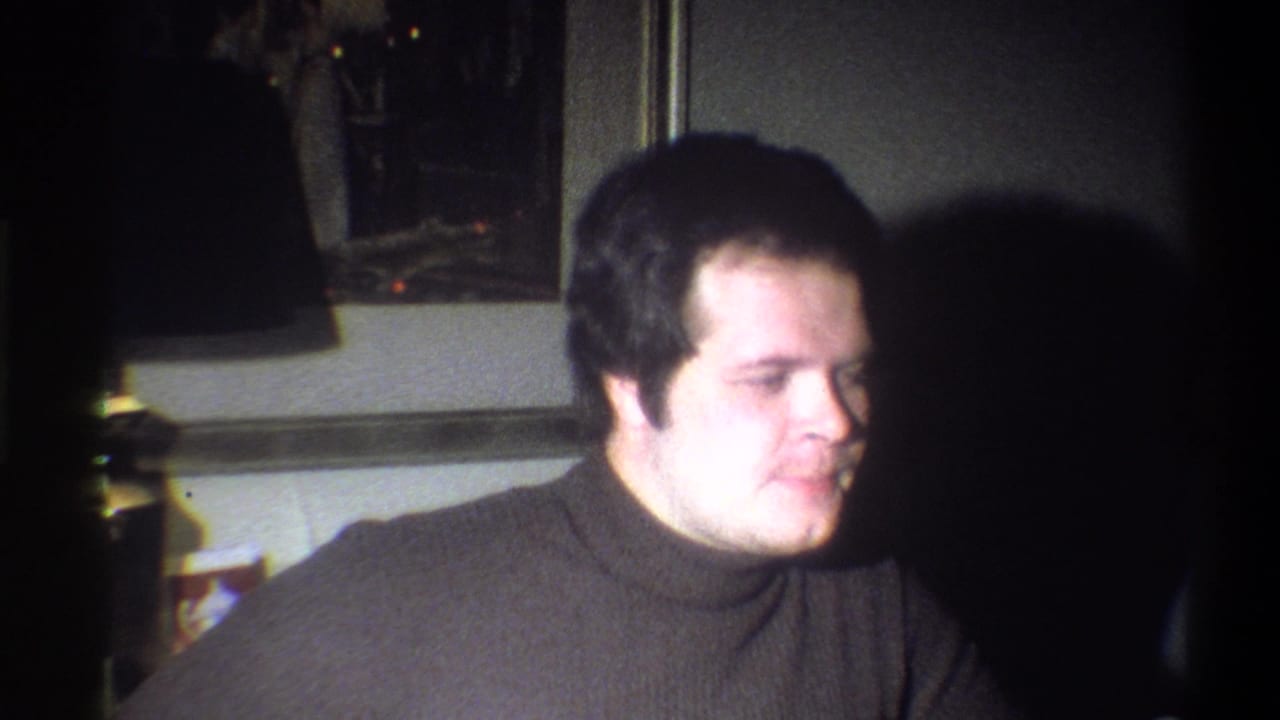 In 1973 Troy, New York home-movie footage, a young man in a dark turtleneck sits indoors with a lamp and framed art behind him.