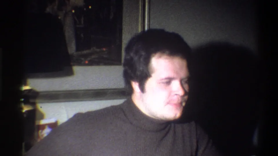 In 1973 Troy, New York home-movie footage, a young man in a dark turtleneck sits indoors with a lamp and framed art behind him.